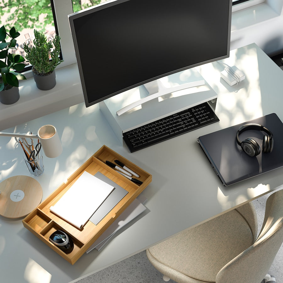 A white monitor stand with a drawer, measuring 10x47x26 cm. Made of durable bamboo with a white aluminum frame, it features compartments for organizing office supplies and a space to store a keyboard. The ergonomic design helps reduce neck and shoulder strain while providing cable management for a tidy workspace.